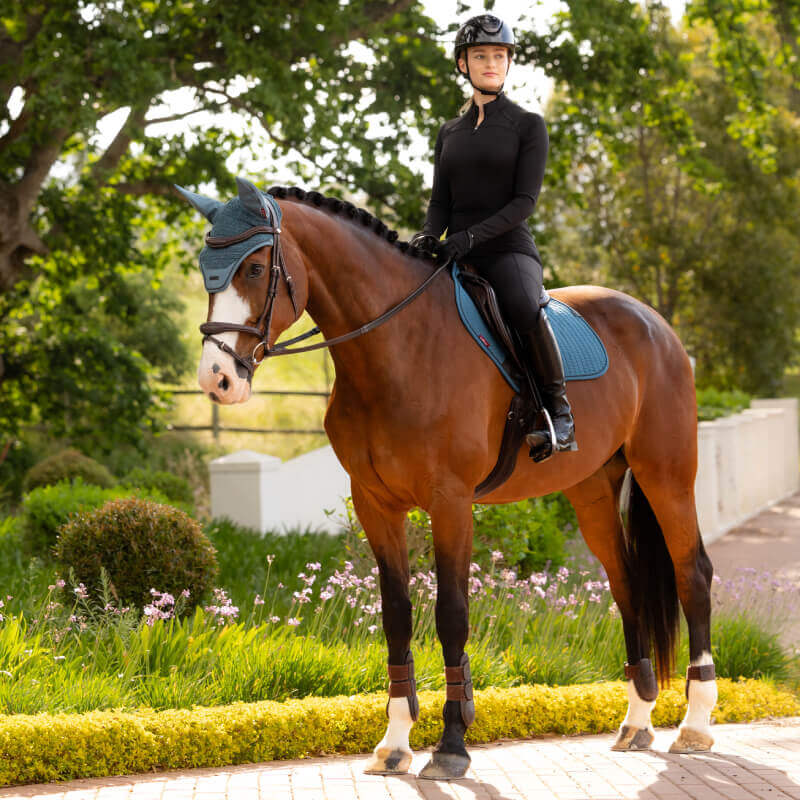 Rider in black sits on a brown horse with a LeMieux Jungle saddle pad in a garden.