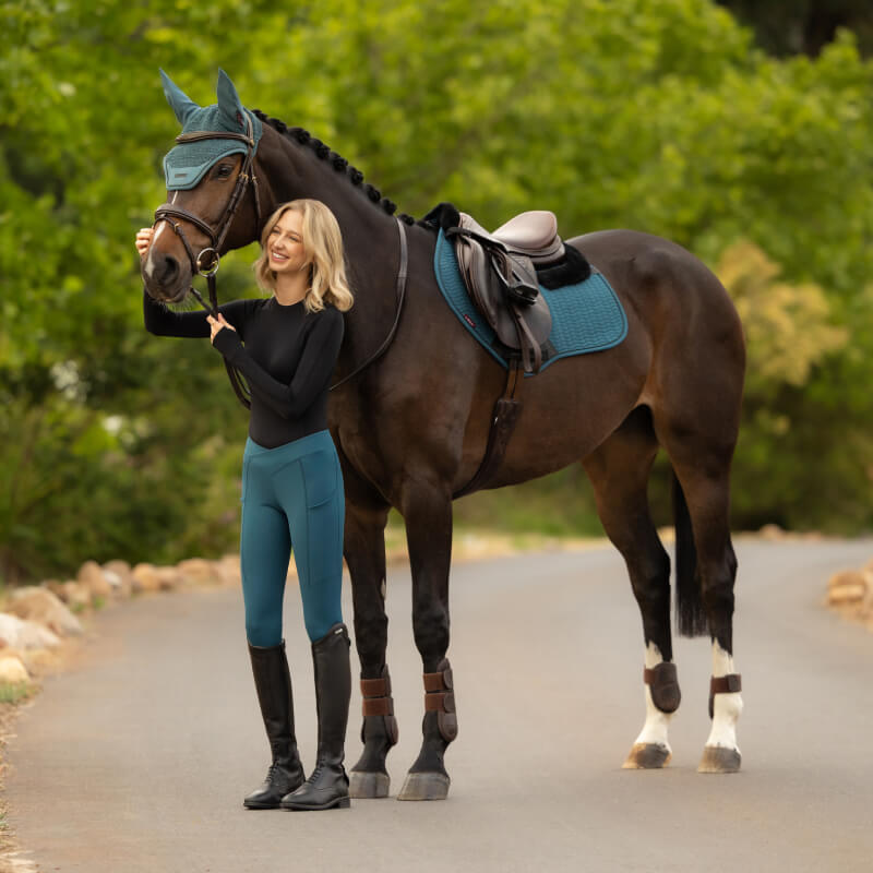 Woman in riding gear stands by a saddled horse with a LeMieux Merino+ Essence Square Jungle pad.