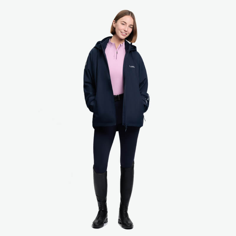 Smiling woman in a LeMieux Indra Waterproof Jacket Navy, pink shirt, black pants, tall boots.