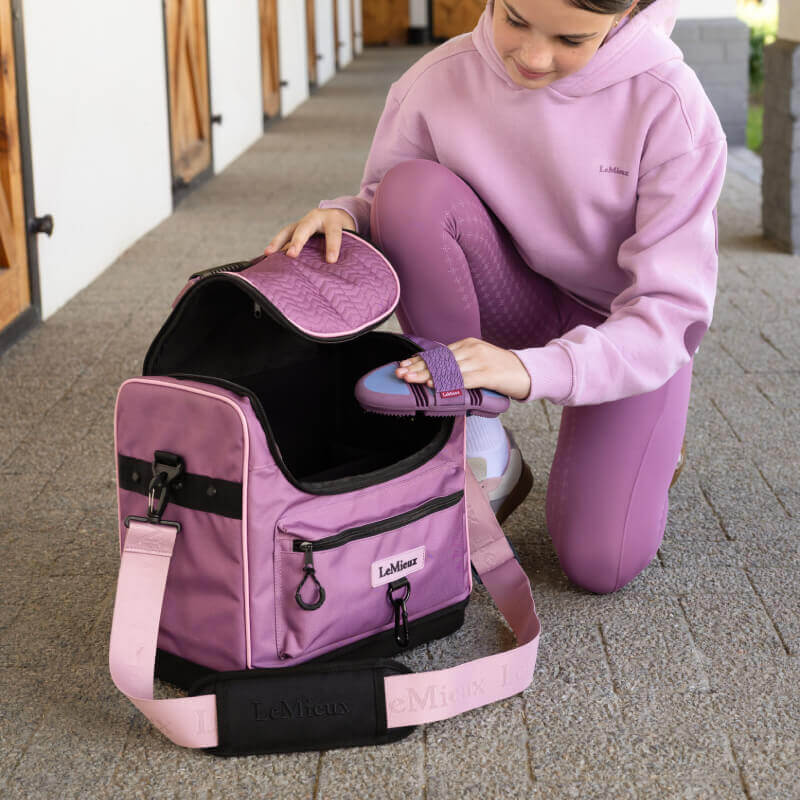 Woman in lilac puts brush into LeMieux Elite Pro Grooming Bag Mallow on walkway.