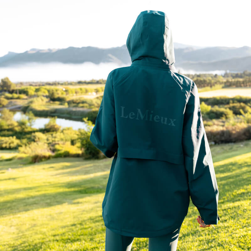 Person in a LeMieux Indra Waterproof Jacket Jungle stands outdoors, facing hills and water.