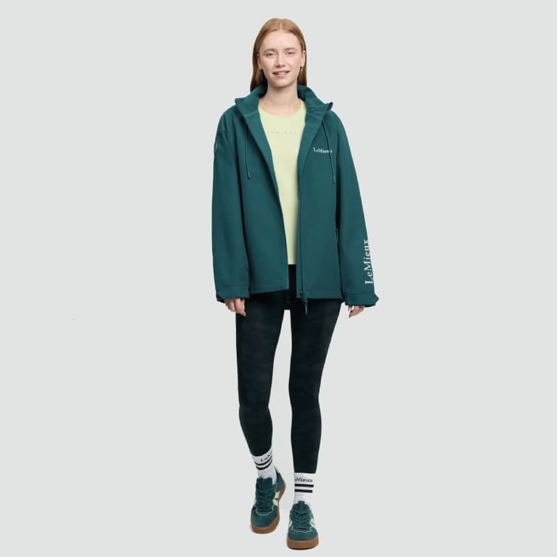 A woman in a LeMieux Indra Waterproof Jacket Jungle, yellow shirt, and black leggings smiles.