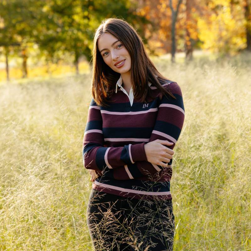 Woman in a LeMieux Sloane Stripe Rugby Shirt Bramble stands in grass with sunlit trees behind.
