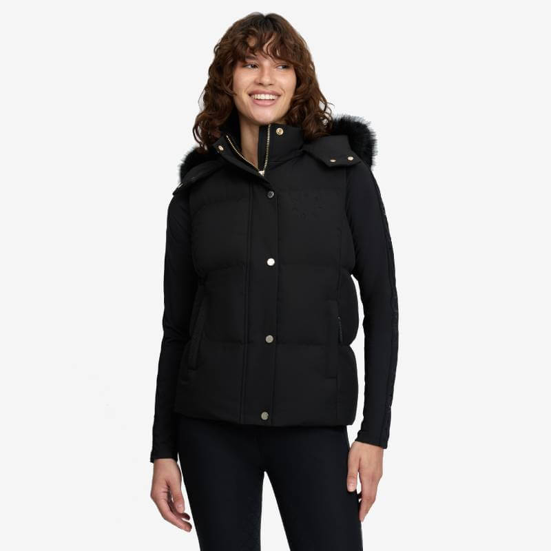 Smiling woman in LeMieux Margot Hooded Gilet Black with fur-trimmed hood, standing.