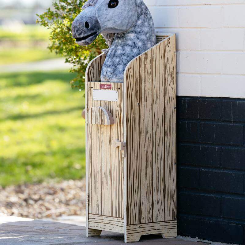Plush horse peeks from LeMieux Hobby Horse Stable Wooden by a white and black wall outdoors.