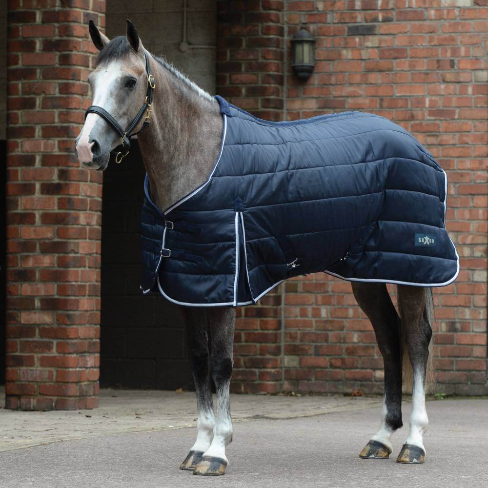 A horse in a Saxon Channel Quilt Stable Rug Medium Navy/White stands by a brick building.