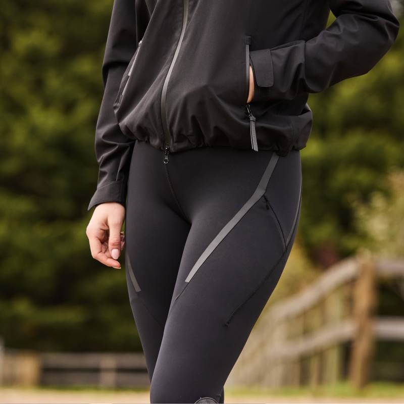 Person wears Weatherbeeta Toulon Lifestyle Tights Black and a jacket, standing by a fence outdoors.