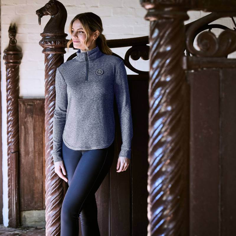 Woman in Weatherbeeta London Layer Long Sleeve Top stands by ornate horse stalls, smiling.