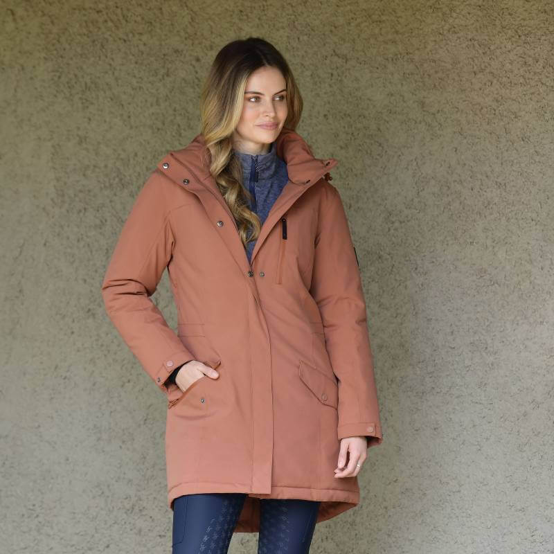 Woman wears Weatherbeeta Kyla II Redwood waterproof jacket, standing by a beige wall.