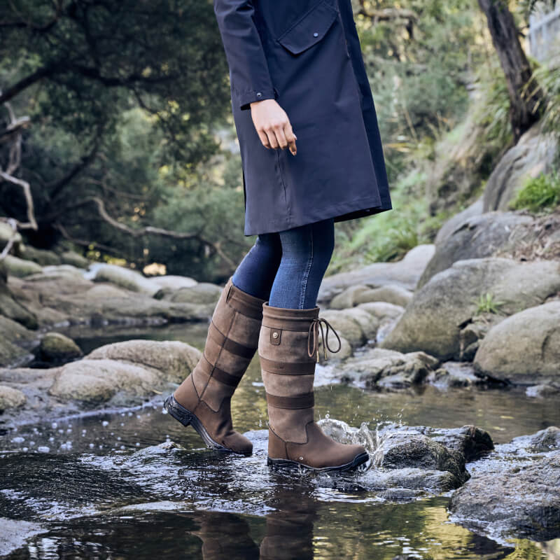 Person in Dublin River Boots IV Chocolate by Dublin, wearing dark coat, walks on rocks in nature.