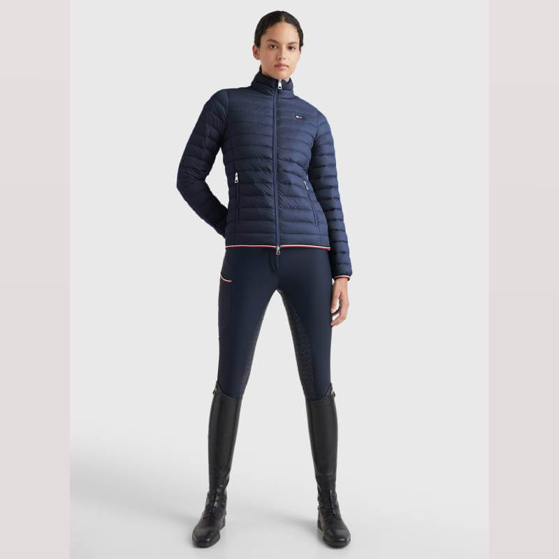 Woman in a Tommy Hilfiger Light Re-Down Jacket, riding pants, and black knee-high boots stands tall.
