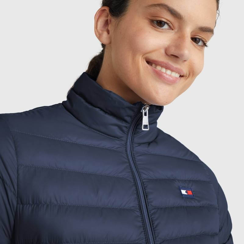 Woman smiling in a Tommy Hilfiger Women Light Re-Down Jacket Desert Sky with chest flag logo.