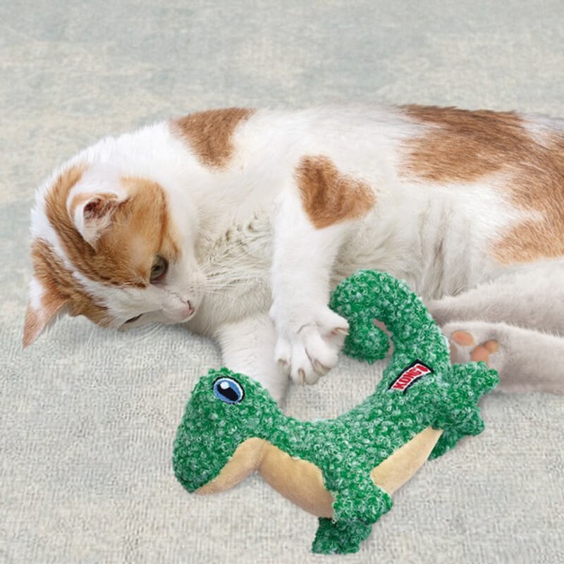 A white and orange cat plays with a green Kong KONG Cat Luvs Lizard toy on a light surface.