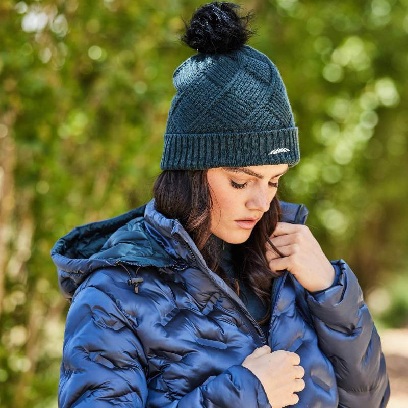 Woman wearing Weatherbeeta Beanie Pine One Size, blue puffer, zipping up outdoors amidst greenery.