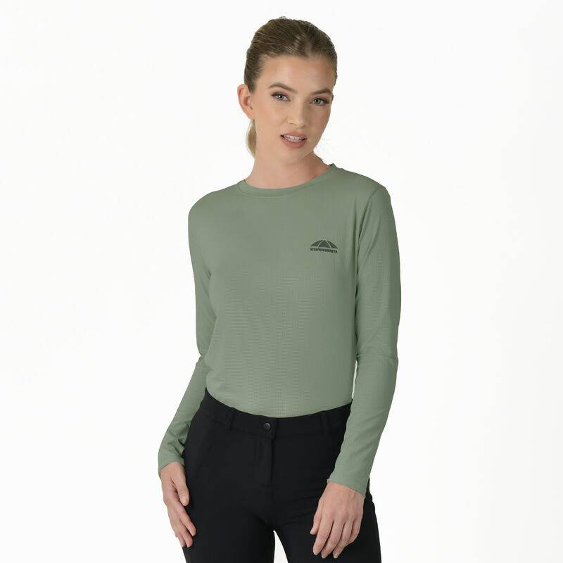 Woman in a Weatherbeeta Relaxed UV Long Sleeve Tee Olive Leaf and black pants stands by a white wall.