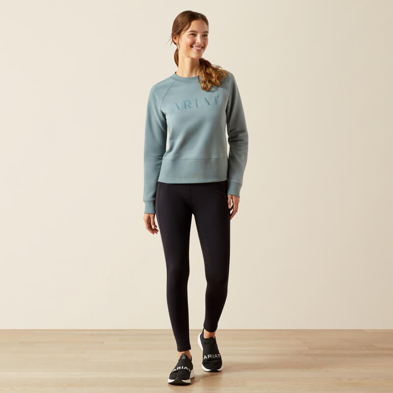 Woman smiling in an Ariat Womens Martine Sweatshirt Steel Sage and black leggings on wood floor.