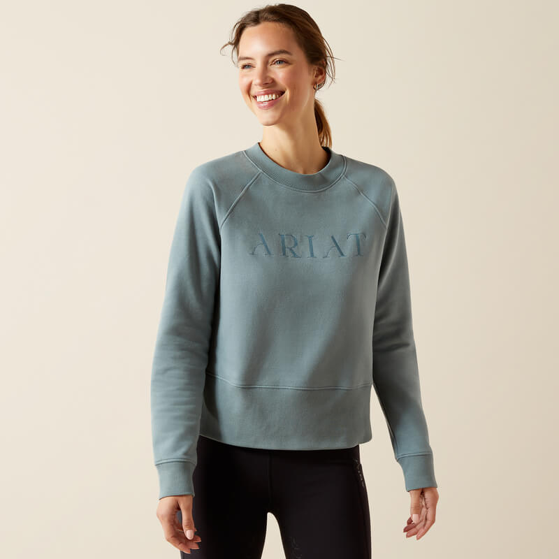 Smiling woman in an Ariat Womens Martine Sweatshirt Steel Sage and black pants stands against beige.