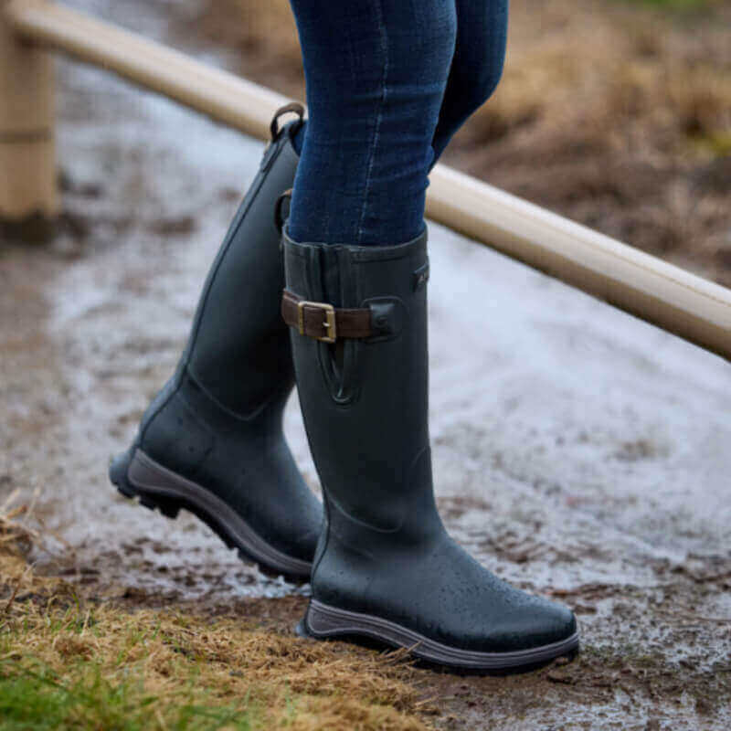 Person wearing tall deep navy Ariat Woodstock boots standing on a muddy path outdoors.