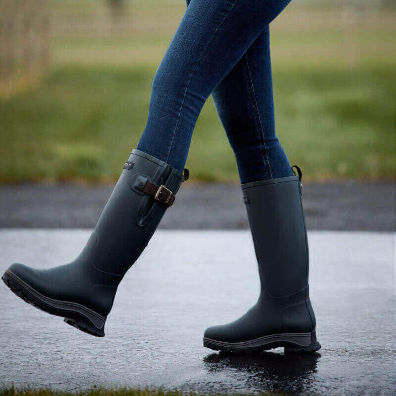 Person in blue jeans wearing Ariat Woodstock Rubber Boot Deep Navy walking on wet pavement.