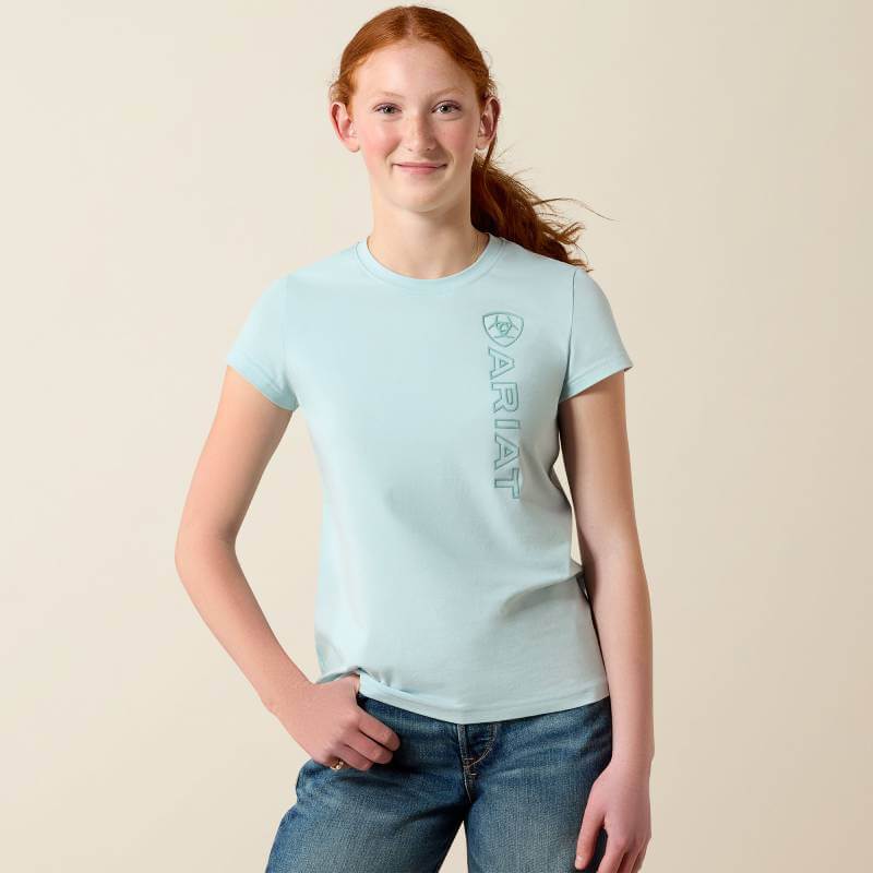 Girl in Ariat Youth Vertical Logo T-Shirt Starlight Blue/Blue Surf stands smiling, hand on hip.