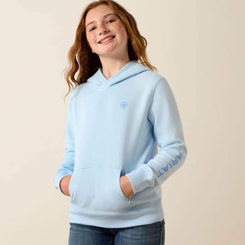 Smiling girl in Ariat Girls 2.0 Hoodie Nantucket Breeze, hands in pockets, beige background.