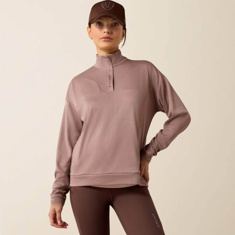 Woman in Ariat Breathe 1/2 Zip Pullover Antler, matching hat, and brown leggings stands confidently.