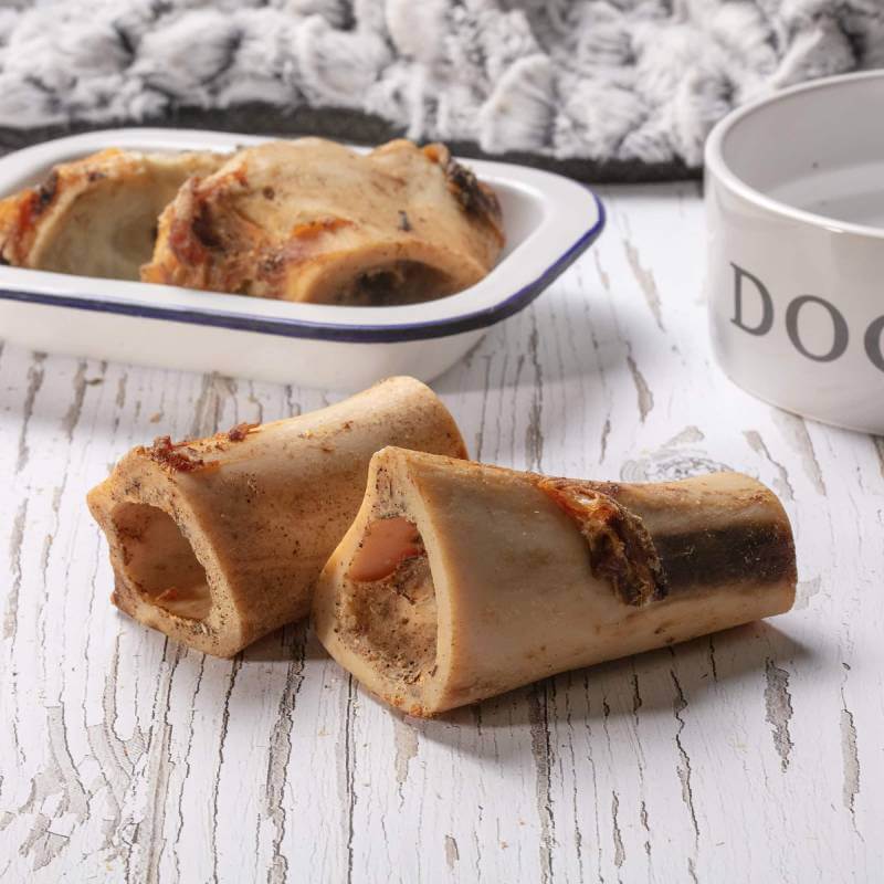 Two Hollings Mini Roast Bone Beef Singles on a white surface, with dog bowl and dish behind.