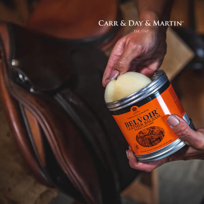 Hand holding Belvoir Leather Balsam Intensive Conditioner 500ml near a brown leather saddle.