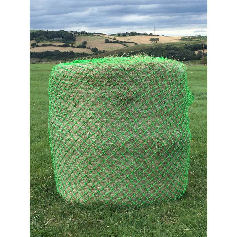 An Elico Wild Boar Bale Net Large Green holds a hay bale on grass with hills in the background.