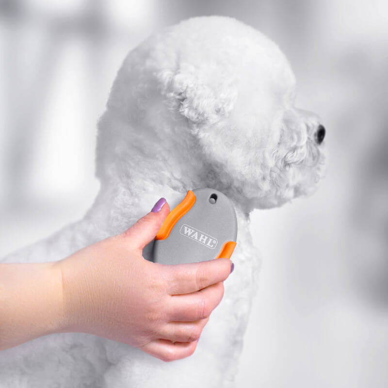 A hand grooming a fluffy white dog with a Wahl Palm Pal Brush Orange Grey to remove loose hair.