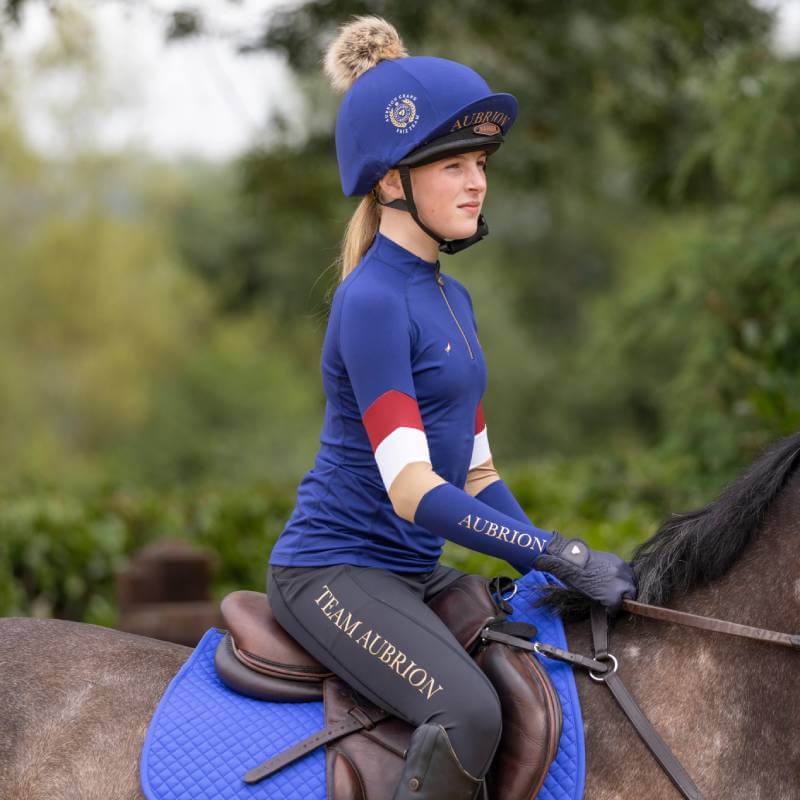 Young rider wears Shires Aubrion Team Long Sleeve Base Layer Navy riding a horse outdoors.