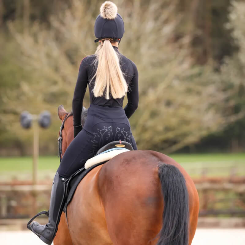 A rider in Aubrion Team Winter Riding Tights Black rides a brown horse outdoors, seen from behind.