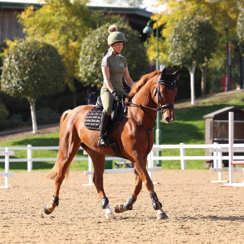 Woman in Aubrion Team Short Sleeve Baselayer Moss rides a brown horse outdoors.