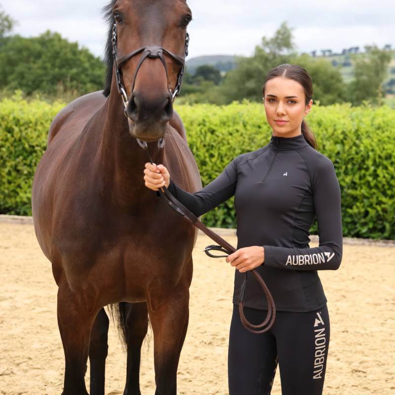 A woman in the Aubrion React Long Sleeve Baselayer YR Shadow stands by a brown horse on sand.