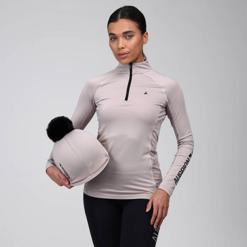 Woman wearing the Aubrion React Long Sleeve Baselayer Sand, holding a beige hat with black pom-pom.