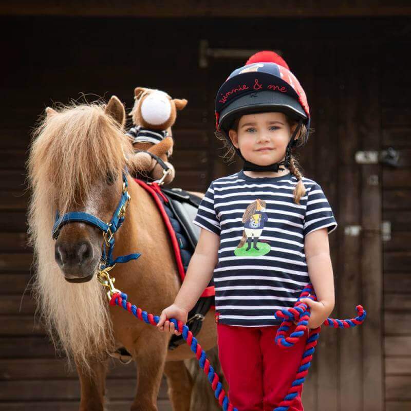 A young girl in a helmet stands beside a small pony, wearing a Shires Winnie & Me Kids T-Shirt.