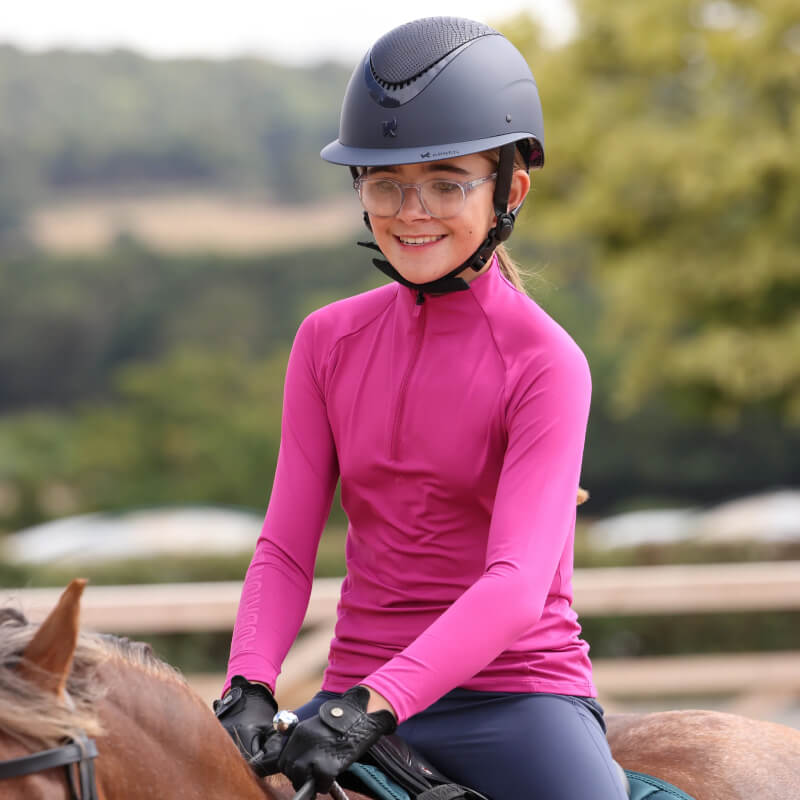 A smiling girl in an Aubrion React Young Rider Fuchsia baselayer and helmet rides a horse outside.