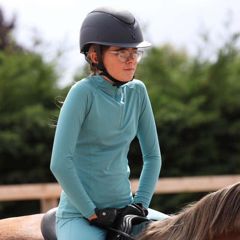 Young rider in Aubrion React Haze Long Sleeve Baselayer and helmet sitting on a horse outdoors.