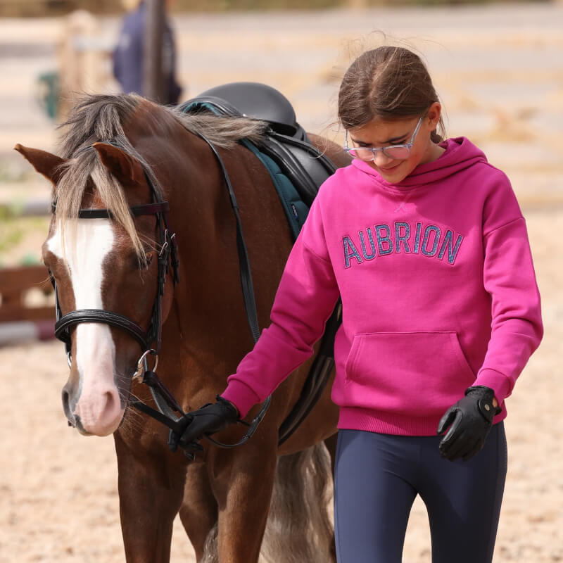 A girl in an Aubrion React Hoodie Young Rider Fuchsia leads a brown horse with a white blaze.