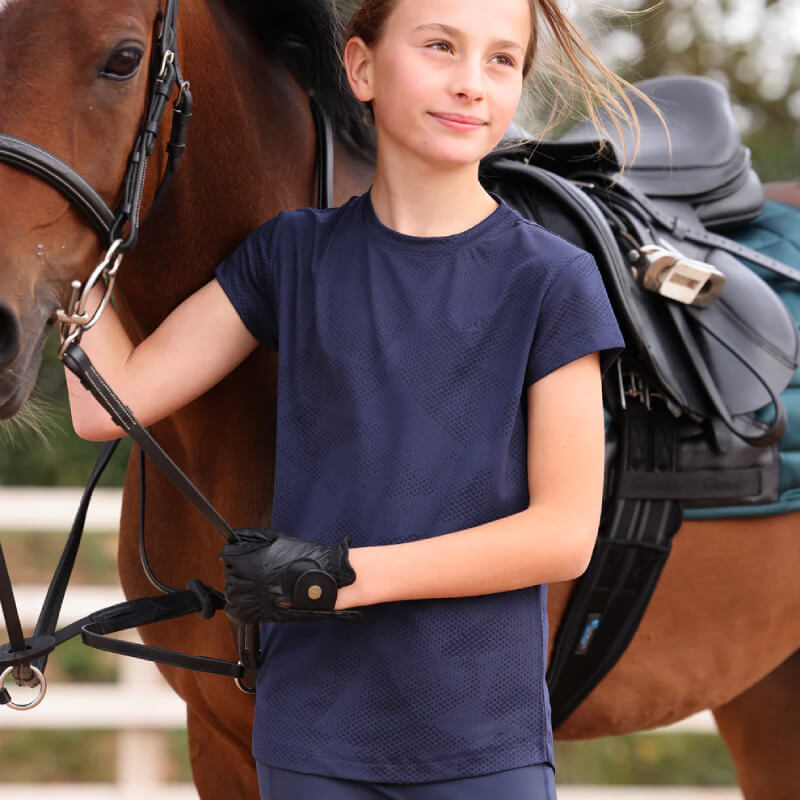 Girl wears Aubrion React Young Rider Tech T-Shirt Midnight beside saddled brown horse outdoors.