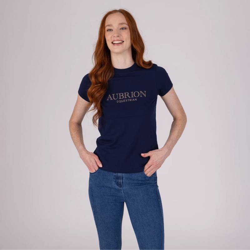 Woman in Aubrion Equestrian Jeans Denim and navy tee smiles, standing against grey.