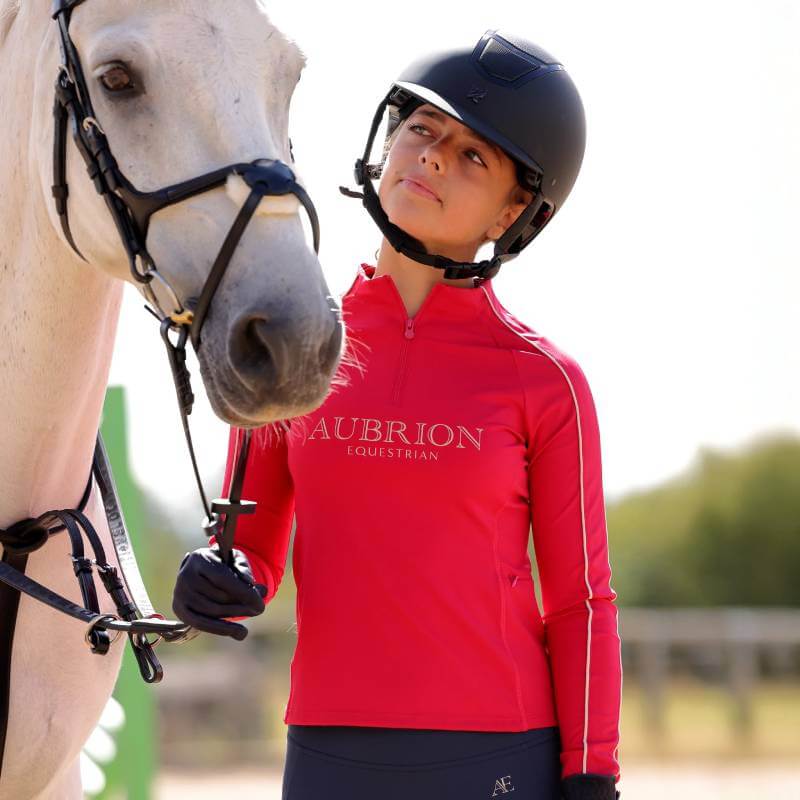 Young rider in Aubrion Dewberry baselayer stands beside a white horse outdoors.