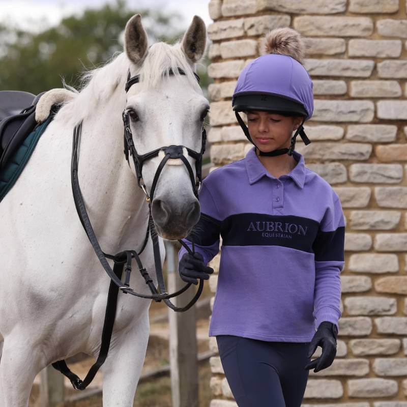 A young rider in an Aubrion sweatshirt leads a white horse by the reins near a stone wall.