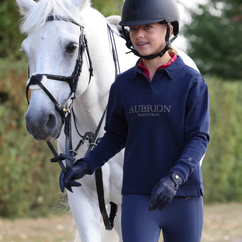 A girl in an Aubrion Equestrian Button Sweatshirt walks beside a white horse outdoors.