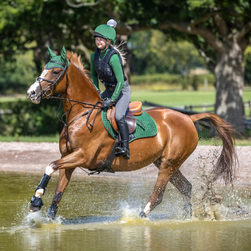 A person rides a brown horse through water with a LeMieux Suede Close Contact Square Hunter Green.