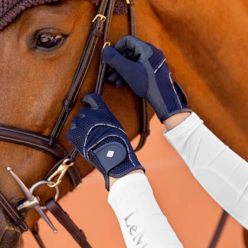 Wearing LeMieux 3D Mesh Riding Gloves Navy, a person adjusts a horse’s bridle on orange.