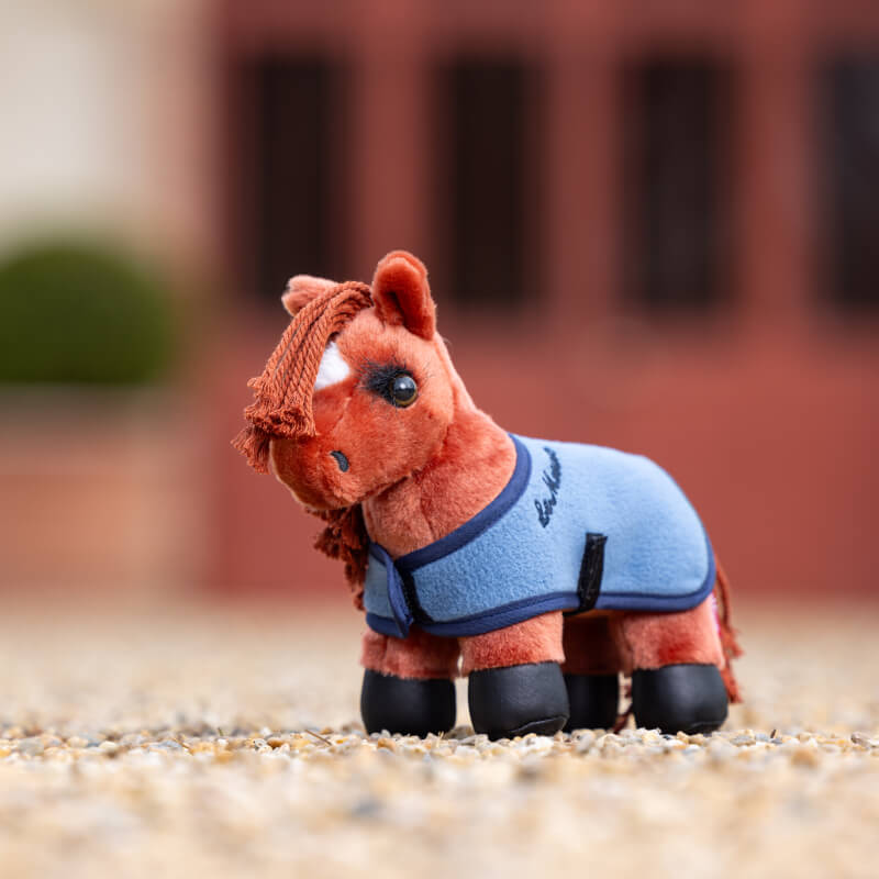 The LeMieux Tiny Pony Thomas is a brown plush pony in a blue jacket and black booties on gravel.
