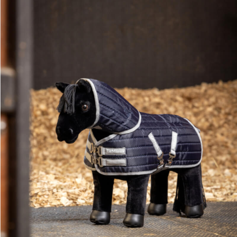 A plush toy horse wears a LeMieux Toy Pony Stable-Tek Rug Navy in a stable with wood shavings.