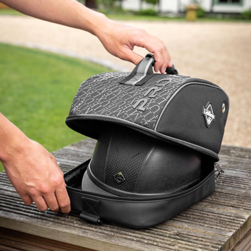 Hands opening a LeMieux Elite Pro Hat Bag Black with a helmet inside on an outdoor wooden surface.