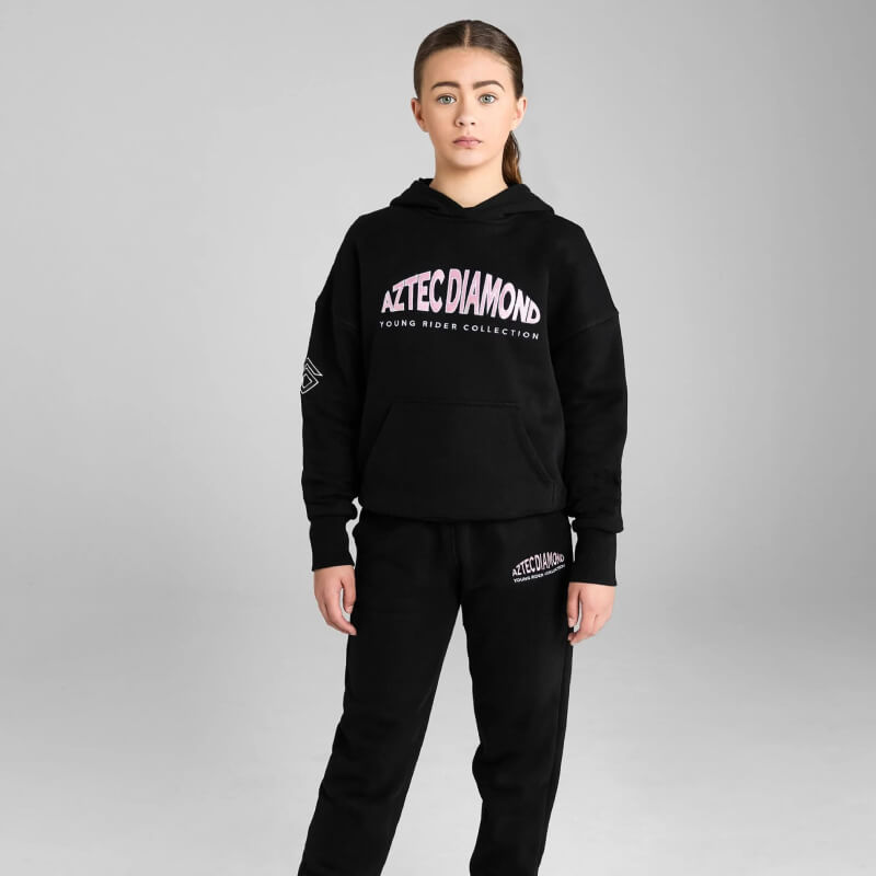 Girl wears Aztec Diamond YR Pull On Hoodie Black/Pink, stands forward by gray background.